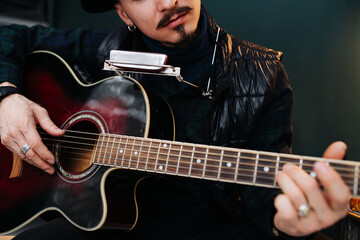 Close up image of a middle aged man in a hat sitting on a stool, plaing guitar at home. He is wearing casual clothes and leather jacket atop.