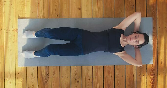 Athletic lady in tracksuit and sneakers does crunches lying on mat on wooden floor at bright sunlight upper view slow motion