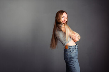 Long-haired blonde in jeans against the background of a gray wall hugs herself and smiles sincerely