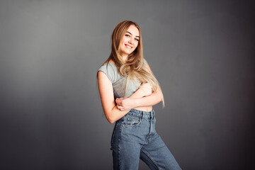 Long-haired blonde in jeans against the background of a gray wall hugs herself and smiles sincerely