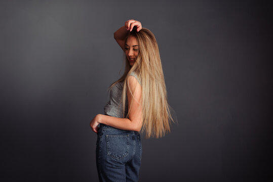 European girl in a gray top and jeans with blond long hair, stands sideways and holds her hand behind her head, looks directly at the wall