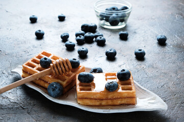 Waffles are laid out on plate, decorated with blueberries, doused with honey from honey spoon.