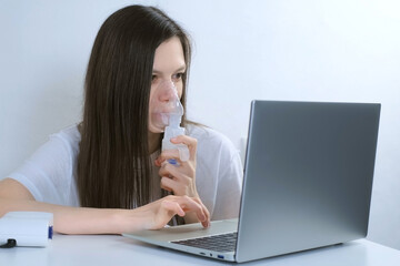 Sick young woman is using nebulizer and inhaler for treatment at home and working on laptop. She is...