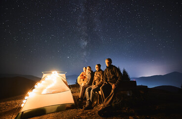 Hikers sitting on bench near illuminated camp tent under blue night sky with stars. Group of...