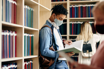 Black university student with face mask reading from a book while learning in library.