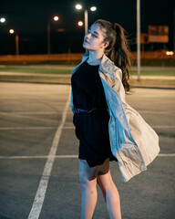 Fototapeta premium Girl in a coat in the parking lot against the backdrop of the night city with lanterns