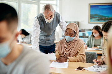 Mature teacher assisting Muslim female student with lecture while wearing face masks in the...
