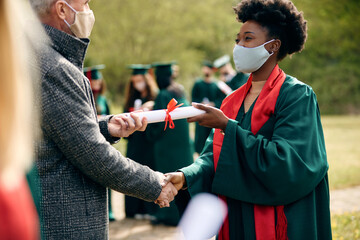 Happy Black student receiving diploma from her professor on graduation day during coronavirus...