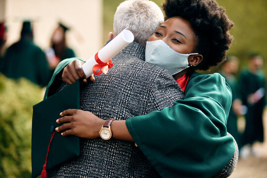 Happy Black student embracing her father after receiving graduation diploma during coronavirus pandemic.