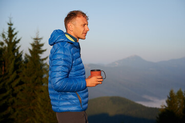 Side view of young man with cup in his hand, squinting looking far away. On the background light blue sky and outlines of mountain peaks in the morning. Concept of hiking, travelling and nature.