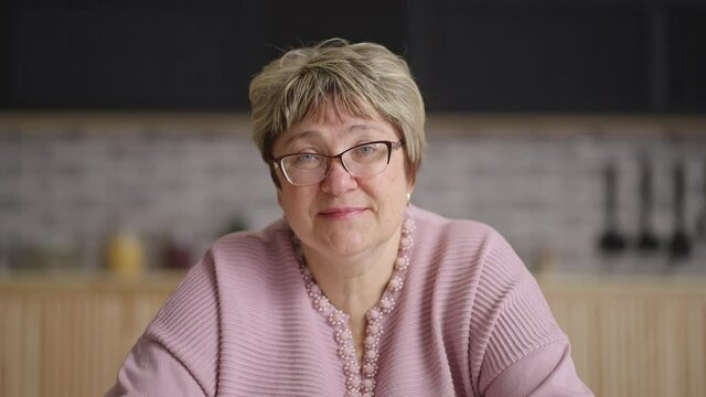 elderly woman is sitting at her home kitchen and looking at camera, listening and nodding, online video chat concept