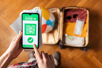 Female's hands hold a passport, protective masks and a smartphone with the app with an immune digital health passport. In the background, a suitcase with clothes in a blur. Top view