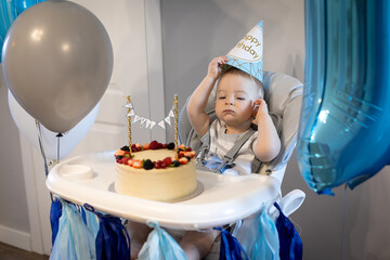 Caucasian baby boy celebrating first birthday at home. Birthday berry cake. Blue decoration, balloon