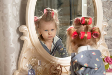 Two-year-old girl. A little girl in curlers looks in the mirror at the dressing table. Party preparation.