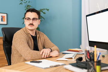 A young handsome smiling man with a moustache is sitting at his desk looking out the window.