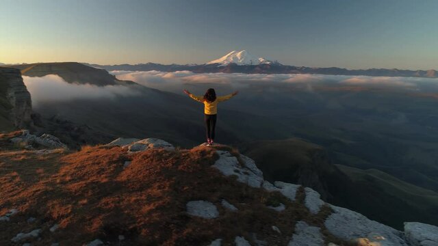 Girl hands spread stands on top of mountain looks at Mount Elbrus. Caucasus emotional inspirational achivement. Travel film Russia. Orange sunset. Scenic clouds fog below. Bermamyt plateau 4k