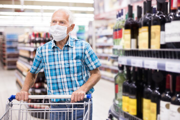 mature european man wearing mask with covid protection chooses bottle of alcohol in supermarket