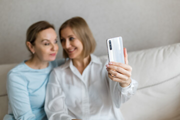Two women sit on the couch and take a selfie on the phone while talking.