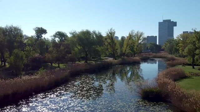 A drone flies over a lake within a natural Parkland with a bridge, trees, and reeds toward a modern business building. Flight on a beautiful spring morning in sunlight with a skyscraper in the back.