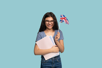 Positive Indian teen exchange student with notebooks holding UK flag, looking at camera and smiling on blue background