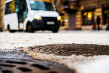 Snowy winter in the big city, the van goes on the road and pedestrians walk on the sidewalk. Close up view of a hatches at the level of the asphalt