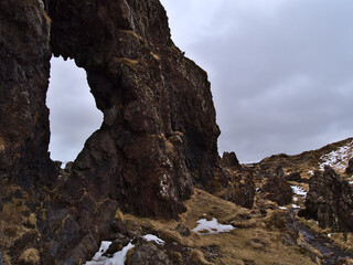 Volcanic rock formations (natural arch) at Djúpalónssandur beach on the western coast of Snæfellsnes peninsula, Iceland in winter season with snow, brown grass and cloudy sky.