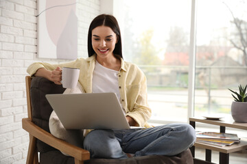 Beautiful young woman with cup of drink using laptop at home
