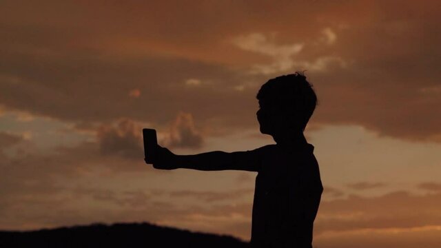 Silhouette of an Indian kid using mobile phone in front of the orange clouds during sunset