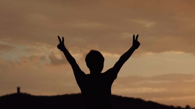 Silhouette of a kid raising his arms and doing peace sign with his hand in front of the beautiful sunset