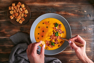 Woman's hands hold crouton and spoon with traditional pumpkin puree cream soup with carrots,...