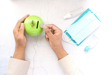 young man saving coins in a jar with face mask and hand sanitizer on table 