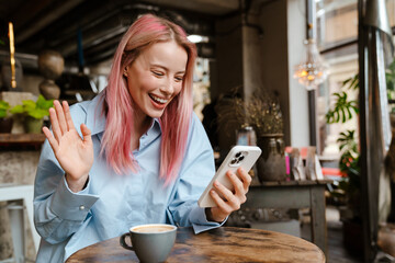 Young smiling woman waving hand and using mobile phone in cafe