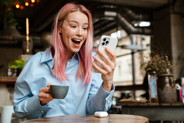 Young excited woman using mobile phone while drinking coffee in cafe