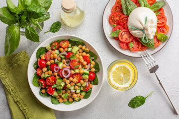 Fresh summer  dinner: mediterranean salad with chickpea, tomatoes, cucumbers, spinach and caprese salad with burrata cheese, basil leaves and tomatoes. Water with lemon. Light background, copy space.