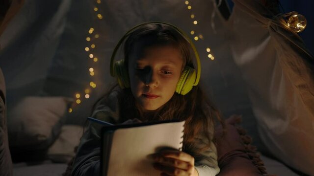 Young girl in headphones thinking and writing while lying on floor in decorative tent at home. Teen listening to music while spending free time in evening. Concept of leisure.