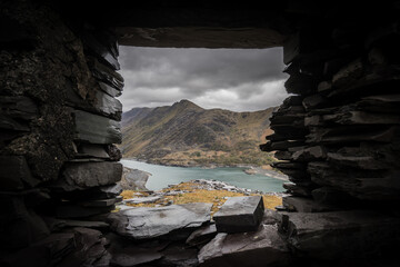 Beautiful view of mount Snowden landscape through window of abandoned miners bothie cottage in Dinorwic slate quarry North Wales. Derelict miners house with blue lake of water and dramatic sky clouds. - obrazy, fototapety, plakaty