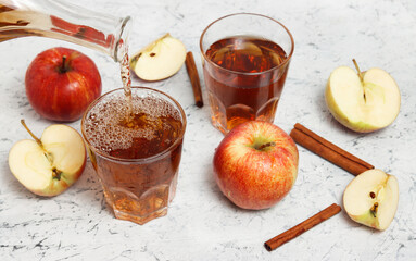 apple juice is pouring from the glass bottle or carafe at the glass full of juice with apples, sliced apples and cinnamon sticks at the background