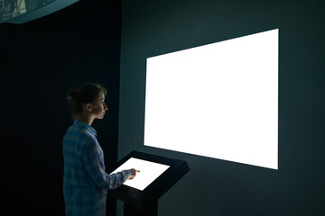 Mock up, futuristic, template and education concept. Woman using electronic kiosk and looking at white blank large interactive wall display in dark room of modern technology museum - mockup image - obrazy, fototapety, plakaty