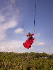 Bali swing trend. Caucasian woman in long red dress swinging in the jungle rainforest. Vacation in Asia. Travel lifestyle. Blue sky. View from back. Bongkasa, Bali, Indonesia