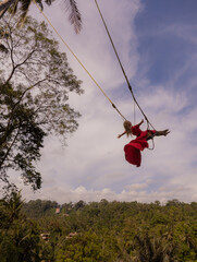 Bali swing trend. Caucasian woman in long red dress swinging in the jungle rainforest. Vacation in Asia. Travel lifestyle. Blue sky. Bongkasa, Bali, Indonesia