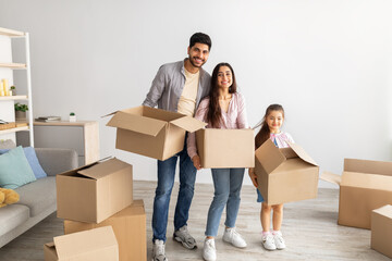 Happy eastern family holding carton boxes and smiling at camera, standing in their new apartment