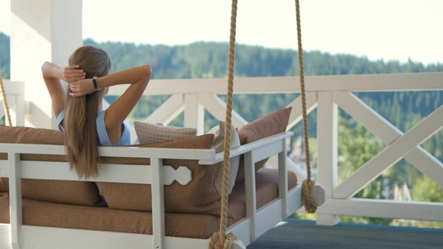 Young happy brunette woman sitting on terrace sofa with soft cushions enjoying warm summer day. Concept of free time on fresh air.