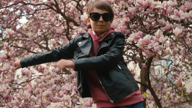 Portrait of a smiling preteen boy in sunglasses dressed in a leather jacket near magnolia tree with flowers. He makes dance moves, rejoices at passing exams. Kids good mood. 