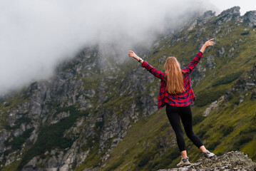 Traveler girl standing raised arms in the top of mountain. Clouds and rocks in the background. Red checkered shirt.