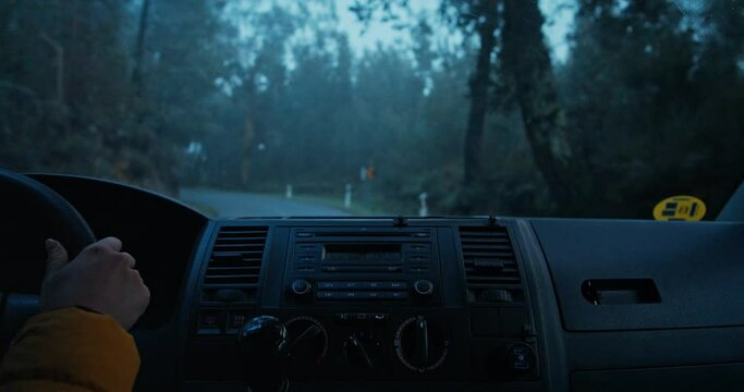 Cinematic POV shot of female driver drive car on moody and foggy dark forest road. Window wipers clean rain drops. Road tripping and roadtrip adventure concept, travel blog