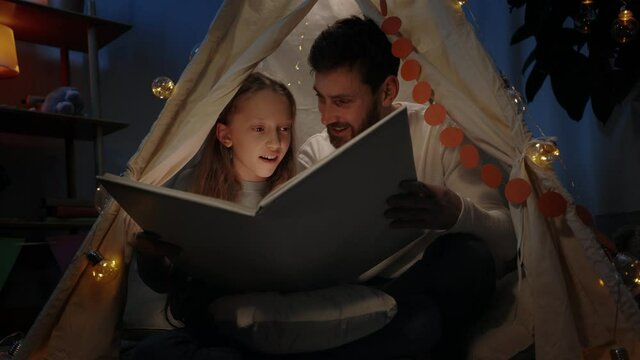 Handsome father and his teen girl opening book together while sitting in decorative makeshift tent . Happy family reading fairy tale while spending good time together. Concept real life.