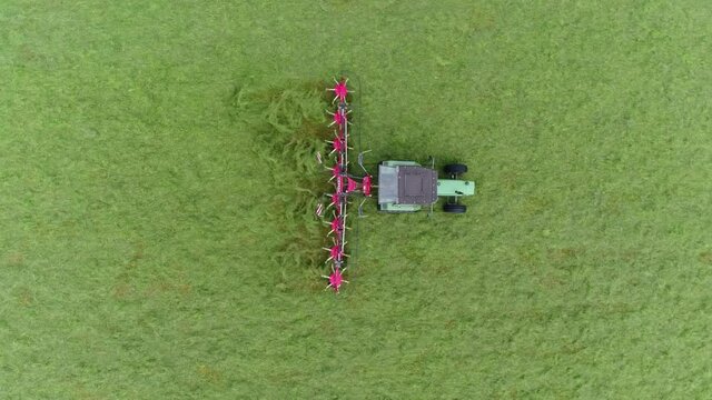 Aerial footage of tractor tedding green meadow mixing up the fresh grass so it can dry and become hay then later it will be picked up and used as animal fodder for example for cows in winter 4k