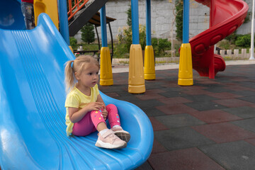 Fototapeta na wymiar Caucasian child sits on a slide after riding, looking forward, colourful playground, happy childhood, summertime. 