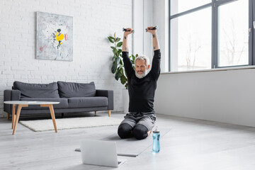 man with grey hair exercising with dumbbells on fitness mat near laptop in living room.