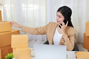 Young asian smart and happy business woman in casual dress working from home with laptop computer and smartphone with online purchase shopping order and box packaging on table (new normal concept)